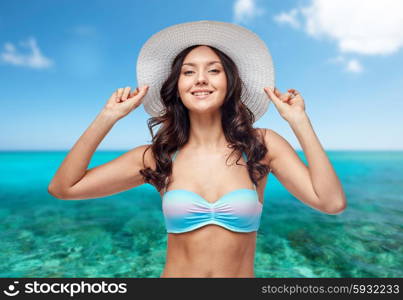 people, summer holidays, travel, tourism and beach concept - happy young woman in bikini swimsuit and sun hat over sea and blue sky background