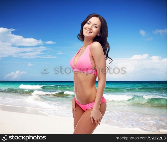 people, summer holidays, travel, tourism and beach concept - happy young woman posing in pink bikini swimsuit over sea shore and blue sky background