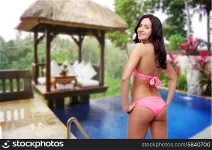 people, summer holidays, travel, tourism and sexual concept - happy young woman in pink bikini swimsuit looking back over hotel resort background