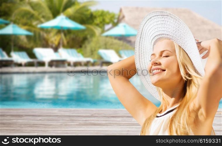 people, summer holidays, travel, tourism and vacation concept - beautiful woman in sun hat enjoying summer over exotic hotel resort beach with swimming pool and sunbeds background