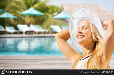 people, summer holidays, travel, tourism and vacation concept - beautiful woman in sun hat enjoying summer over exotic hotel resort beach with swimming pool and sunbeds background