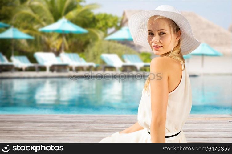 people, summer holidays, travel, tourism and vacation concept - beautiful woman in sun hat enjoying summer outdoors over exotic hotel resort beach with swimming pool and sunbeds background