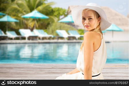 people, summer holidays, travel, tourism and vacation concept - beautiful woman in sun hat enjoying summer outdoors over exotic hotel resort beach with swimming pool and sunbeds background