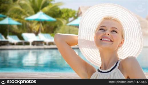 people, summer holidays, travel, tourism and vacation concept - beautiful woman in sun hat enjoying summer outdoors over exotic hotel resort beach with swimming pool and sunbeds background