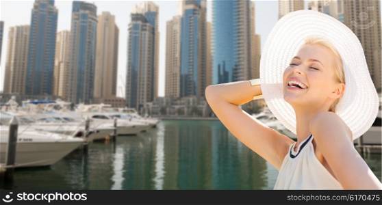 people, summer holidays, travel, tourism and vacation concept - happy beautiful woman in sun hat enjoying summer over dubai city waterfront and boats background