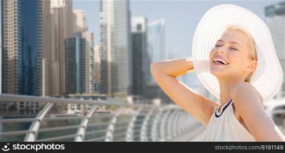 people, summer holidays, travel, tourism and vacation concept - happy beautiful woman in sun hat enjoying summer over dubai city waterfront background