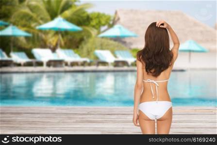 people, summer holidays, travel, tourism and vacation concept - woman in bikini swimsuit from back over exotic hotel resort beach with swimming pool and sunbeds background