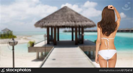 people, summer holidays, travel, tourism and vacation concept - woman in bikini swimsuit from back over maldives beach with bungalow background