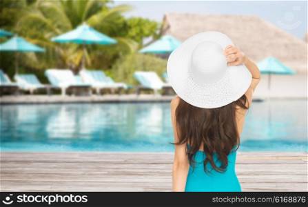 people, summer holidays, travel, tourism and vacation concept - woman in swimsuit and sun hat from back over exotic hotel resort beach with swimming pool and sunbeds background