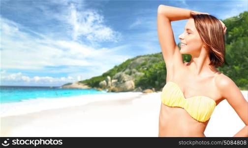 people, summer holidays, vacation and travel concept - happy smiling young woman posing in bikini swimsuit posing with raised hand over exotic tropical beach and sea shore background. happy woman in bikini swimsuit on tropical beach. happy woman in bikini swimsuit on tropical beach