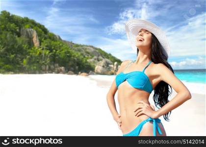 people, summer holidays, vacation and travel concept - happy smiling young woman posing in bikini swimsuit and sun hat posing over exotic tropical beach and sea shore background. happy woman in bikini and sun hat on beach