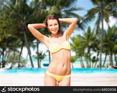 people, summer holidays, vacation and travel concept - happy young woman posing in bikini swimsuit with raised hands over exotic tropical beach with palm trees and pool background