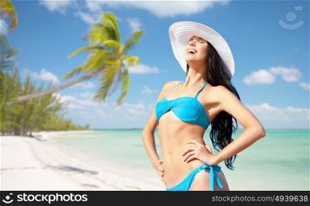 people, summer holidays, vacation and travel concept - happy young woman posing in bikini swimsuit and hat over exotic tropical beach with palm trees and sea shore background. happy woman in bikini swimsuit on tropical beach