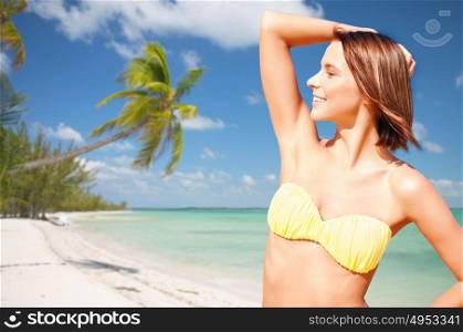 people, summer holidays, vacation and travel concept - happy young woman posing in bikini swimsuit with raised hand over exotic tropical beach with palm trees and sea shore background. happy woman in bikini swimsuit on tropical beach