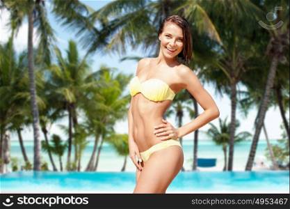 people, summer holidays, vacation and travel concept - happy young woman posing in bikini swimsuit over exotic tropical beach with palm trees and pool background. happy woman in bikini swimsuit on tropical beach