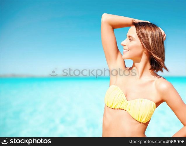 people, summer holidays, vacation and travel concept - happy young woman posing in bikini swimsuit with raised hand on beach over blue sky and sea background. happy woman in bikini swimsuit on tropical beach