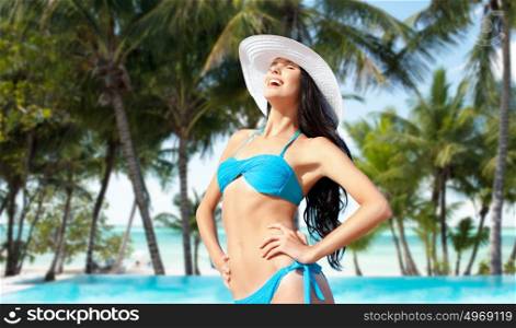 people, summer holidays, vacation and travel concept - happy young woman posing in bikini swimsuit and hat over exotic tropical beach with palm trees and pool background. happy woman in bikini swimsuit on tropical beach