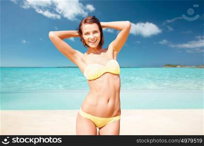 people, summer holidays, vacation and travel concept - happy young woman posing in bikini swimsuit with raised hands over exotic tropical beach background. happy woman in bikini swimsuit on tropical beach