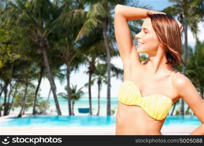 people, summer holidays, vacation and travel concept - happy young woman posing in bikini swimsuit with raised hand over exotic tropical beach with palm trees and pool background. happy woman in bikini swimsuit on tropical beach
