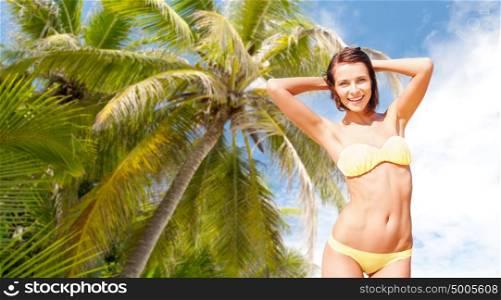people, summer holidays, vacation and travel concept - happy young woman posing in bikini swimsuit with raised hands over palm tree background. happy woman in bikini swimsuit over palm tree