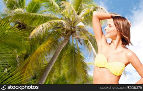people, summer holidays, vacation and travel concept - happy young woman posing in bikini swimsuit with raised hand over palm tree background. happy woman in bikini swimsuit over palm tree