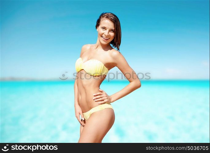 people, summer holidays, vacation and travel concept - happy young woman posing in bikini swimsuit on beach over sea and blue sky background. happy woman in bikini swimsuit on tropical beach