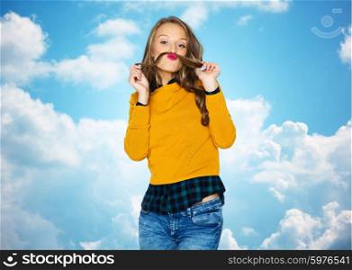 people, summer, style and fashion concept - happy young woman or teen girl in casual clothes having fun making mustache of her hair strand over blue sky and clouds background