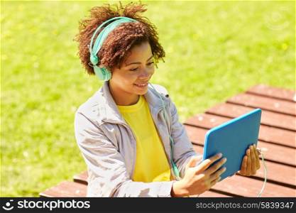 people, summer, technology and leisure concept - happy african american young woman in headphones with tablet pc computer listening to music or watching video outdoors