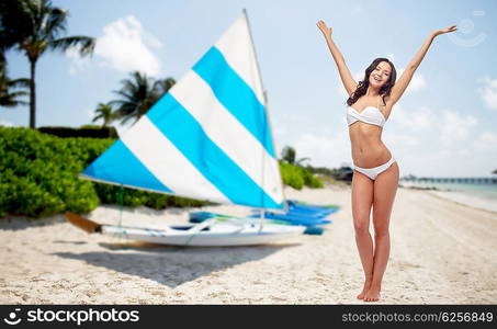 people, summer vacation, tourism and travel concept - happy young woman in white bikini swimsuit posing or dancing with raised hands over sailing boats on tropical beach background