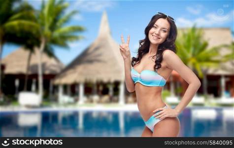 people, summer vacation, travel and gesture concept - happy woman in bikini swimsuit showing victory hand sign over swimming pool, bungalow and palm trees at hotel resort background
