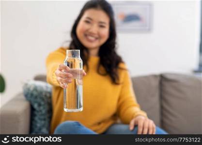 people, sustainability and leisure concept - close up of happy smiling asian young woman in yellow sweater sitting on sofa with glass bottle of water at home. close up of woman with bottle of water at home
