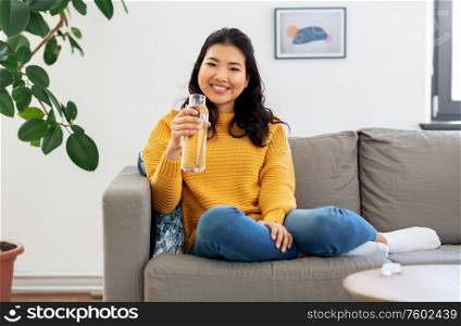 people, sustainability and leisure concept - happy smiling asian young woman in yellow sweater sitting on sofa and drinking water from glass bottle at home. smiling asian young woman drinking water at home