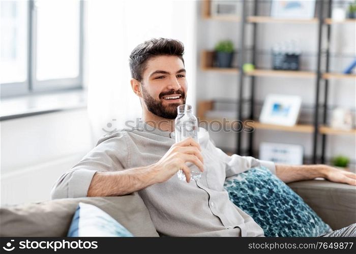 people, sustainability and leisure concept - happy smiling young man sitting on sofa and drinking water from glass bottle at home. happy man drinking water from glass bottle at home