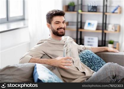 people, sustainability and leisure concept - happy smiling young man sitting on sofa and drinking water from glass bottle at home. happy man drinking water from glass bottle at home