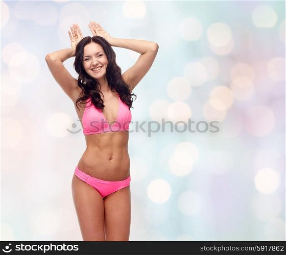 people, swimwear, summer beach and sexual concept - happy young woman in pink bikini swimsuit making bunny ears gesture over blue holidays lights background