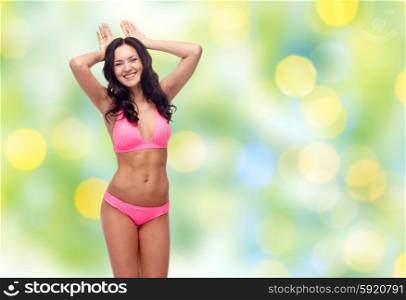 people, swimwear, summer beach and sexual concept - happy young woman in pink bikini swimsuit making bunny ears gesture over green holidays lights background