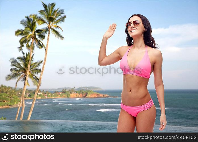 people, swimwear, summer, travel and gesture concept - happy young woman in sunglasses and pink swimsuit waving hand over Sri Lanka beach with palms swimming pool background