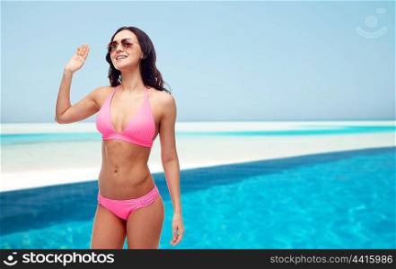 people, swimwear, summer, travel and gesture concept - happy young woman in sunglasses and pink swimsuit waving hand over maldives beach with swimming pool background
