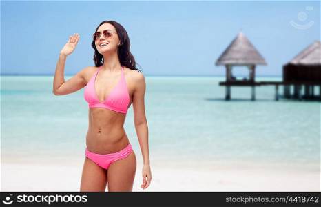 people, swimwear, summer travel and gesture concept - happy young woman in sunglasses and pink swimsuit waving hand over maldives beach with bungalow background