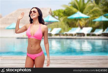 people, swimwear, summer, travel and gesture concept - happy young woman in sunglasses and pink swimsuit waving hand over resort swimming pool with parasols and sunbeds background