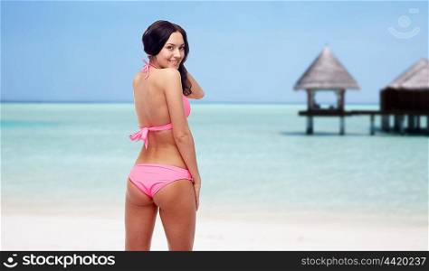 people, swimwear, summer, travel and sexual concept - happy young woman in pink bikini swimsuit looking back over maldives beach with bungalow background