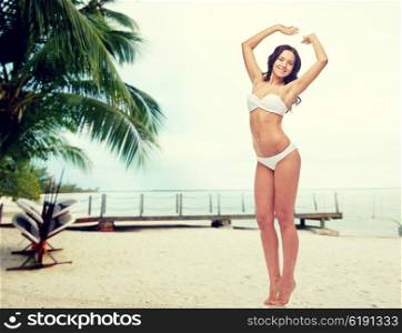 people, swimwear, tourism, travel and summer concept - happy young woman posing in white bikini swimsuit with raised hands over berth on tropical beach background