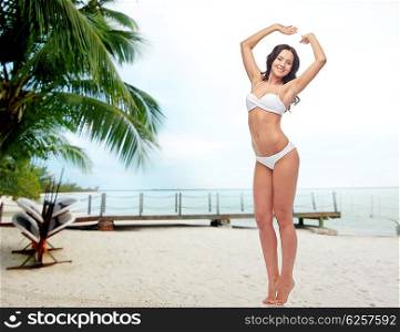 people, swimwear, tourism, travel and summer concept - happy young woman posing in white bikini swimsuit with raised hands over berth on tropical beach background