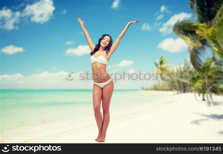 people, swimwear, travel, summer holidays and sea concept - happy young woman in white bikini swimsuit posing or dancing with raised hands over tropical beach background