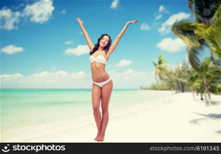 people, swimwear, travel, summer holidays and sea concept - happy young woman in white bikini swimsuit posing or dancing with raised hands over tropical beach background