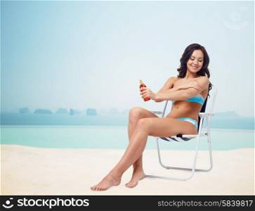 people, tanning, skincare, summer and beach concept - happy young woman in bikini swimsuit sunbathing on folding chair and applying sunscreen to skin over infinity edge pool at hotel resort background