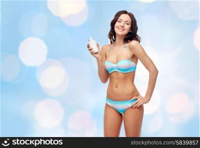 people, tanning, skincare, summer and beach concept - happy young woman in bikini swimsuit holding sunscreen bottle over blue holidays lights background
