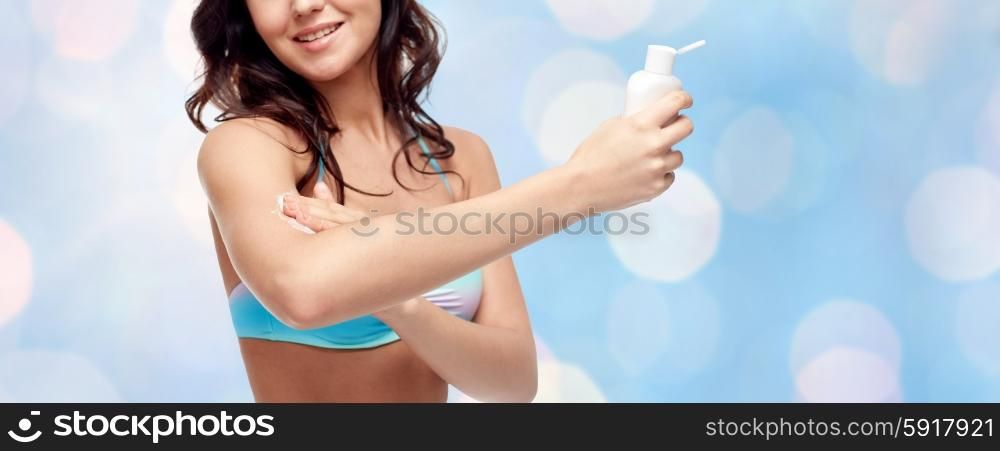 people, tanning, skincare, summer and beach concept - happy young woman in bikini swimsuit holding and applying sunscreen to her arm over blue holidays lights background