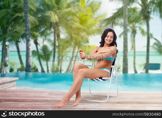 people, tanning, skincare, summer and beach concept - happy young woman in bikini swimsuit sunbathing on folding chair and applying sunscreen to her skin over tropical beach at hotel resort background