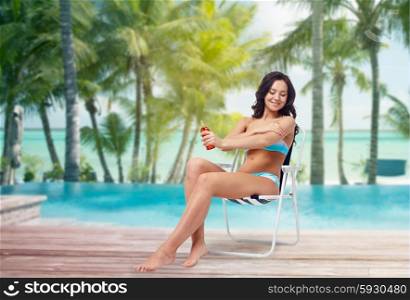 people, tanning, skincare, summer and beach concept - happy young woman in bikini swimsuit sunbathing on folding chair and applying sunscreen to her skin over tropical beach at hotel resort background